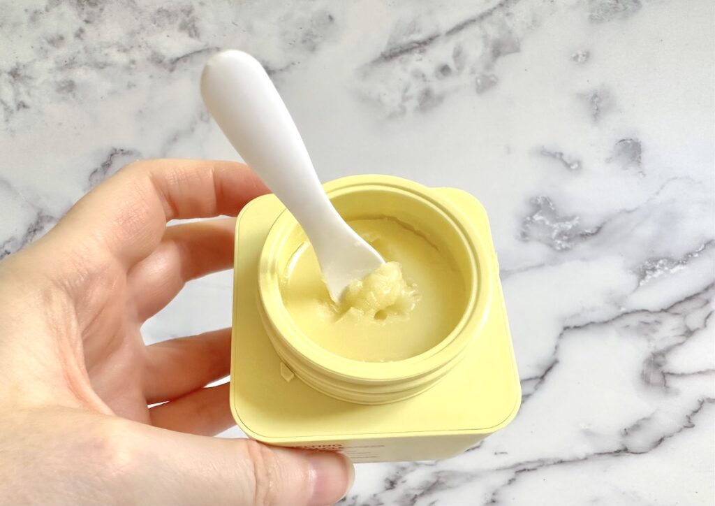 Byoma Melting Balm Cleanser Cleansing Balm, open jar with white cosmetic spatula, handheld.