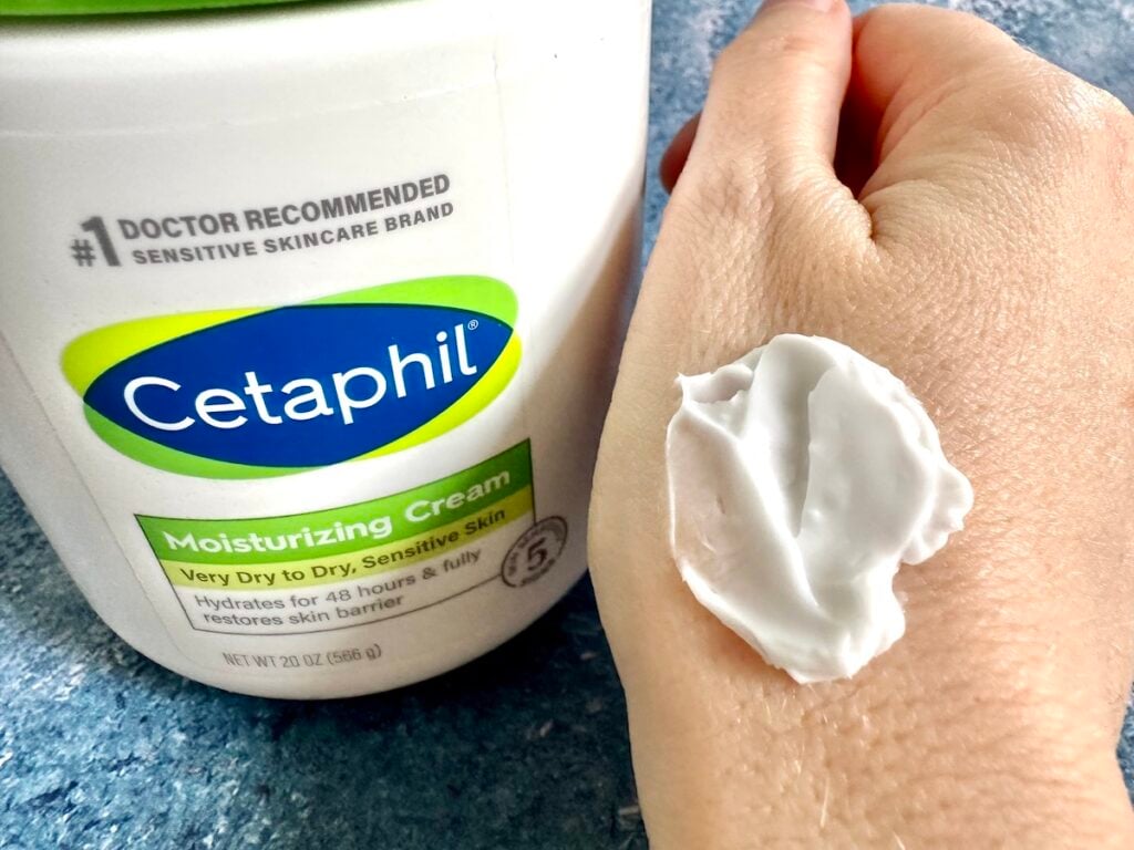 Cetaphil Moisturizing Cream tub next to cream sampled on back of hand.