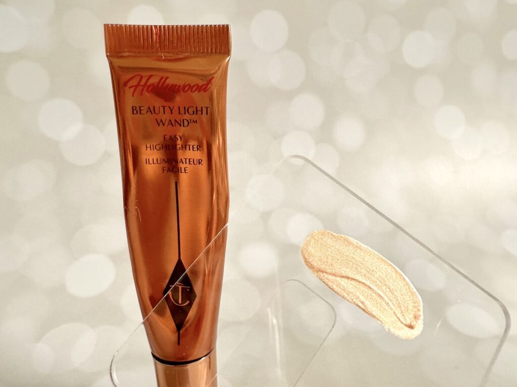 Charlotte Tilbury Hollywood Beauty Light Wand in the shade Spotlight, open with applicator sponge, tube, next to sample on clear cosmetic spatula.