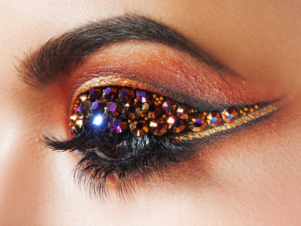 Mosaic of multi-faceted rhinestones in shades of purple, gold, and orange, meticulously placed over a golden eyeshadow base. Winged eye makeup look.