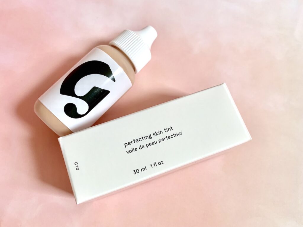 Glossier Perfecting Skin Tint and box flatlay on pink background.