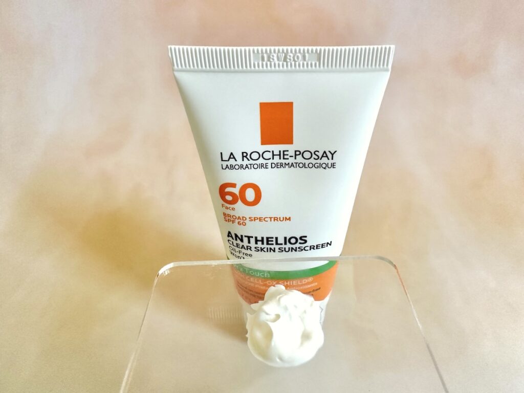 La Roche-Posay Anthelios Clear Skin Sunscreen SPF 60, tube behind sunscreen sampled on clear cosmetic spatula.