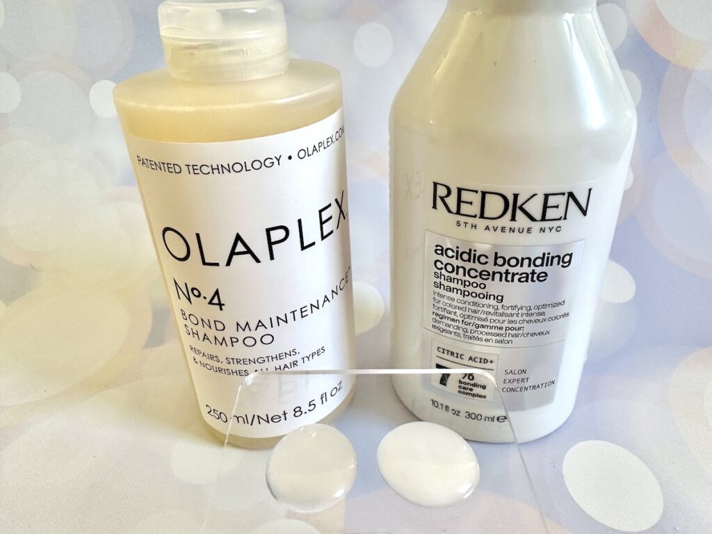 Redken Acidic Bonding Concentrate Shampoo and Olaplex No. 4 Bond Maintenance Shampoo bottles behind samples on clear plastic spatula.