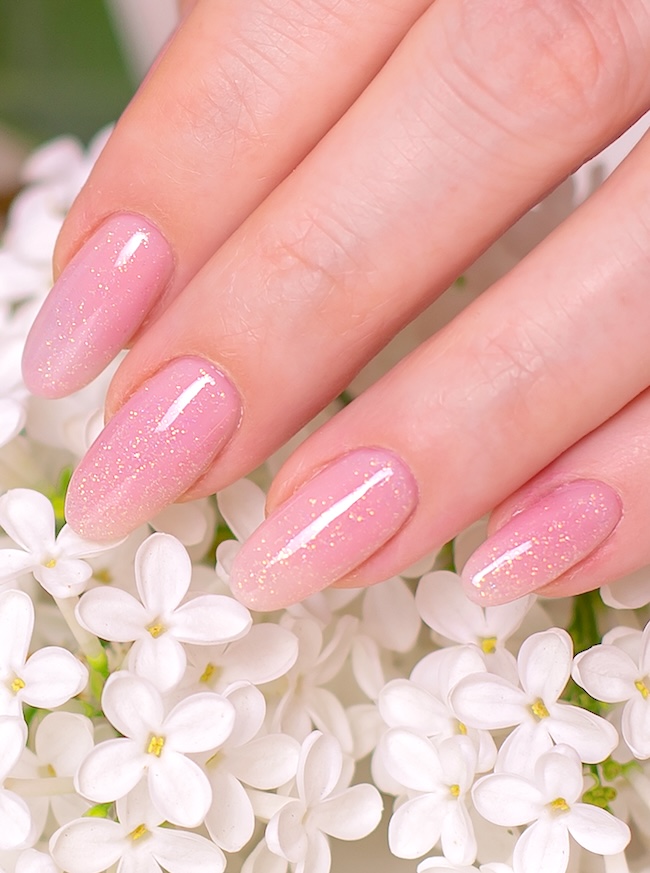 Golden glitter on oval pink nails touching white flowers.