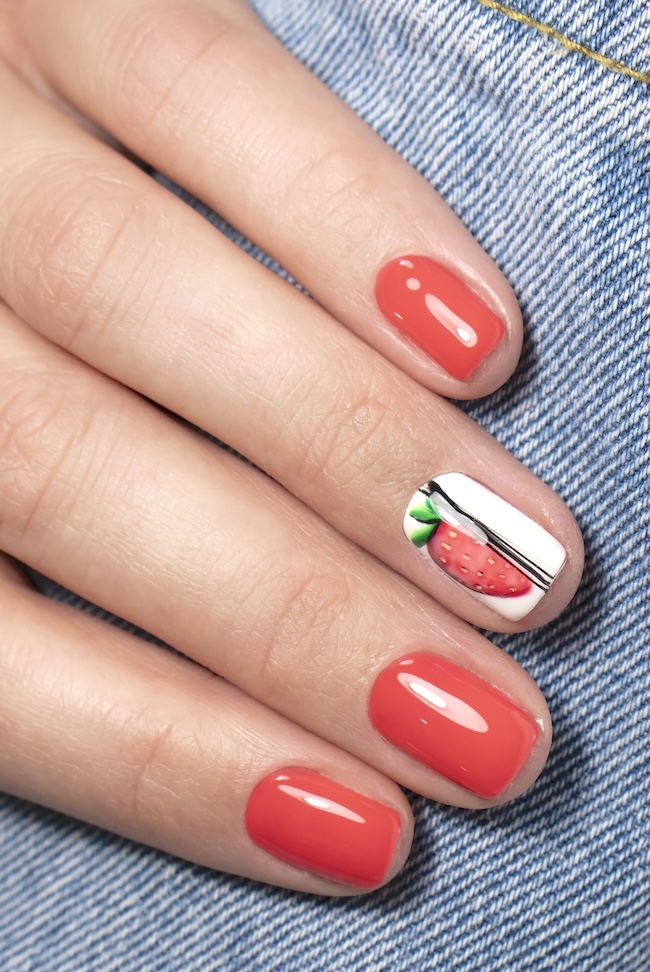 Short coral red nails with strawberry nail art on denim background.