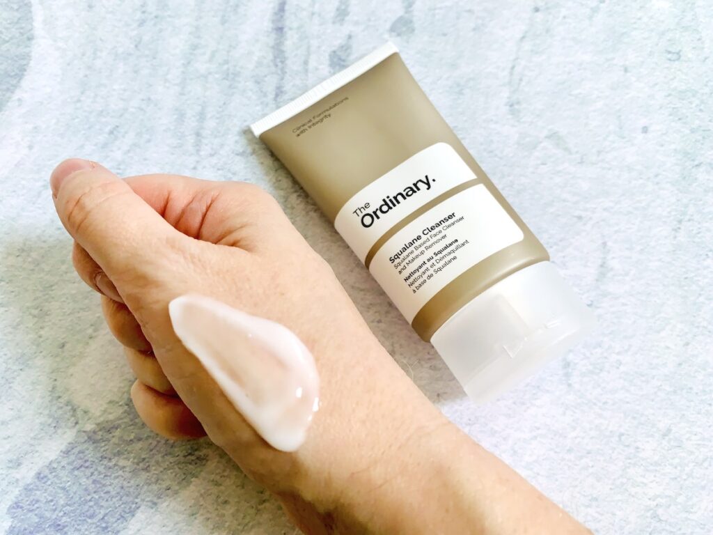 The Ordinary Squalane Cleanser tube and sampled on hand on a blue background.