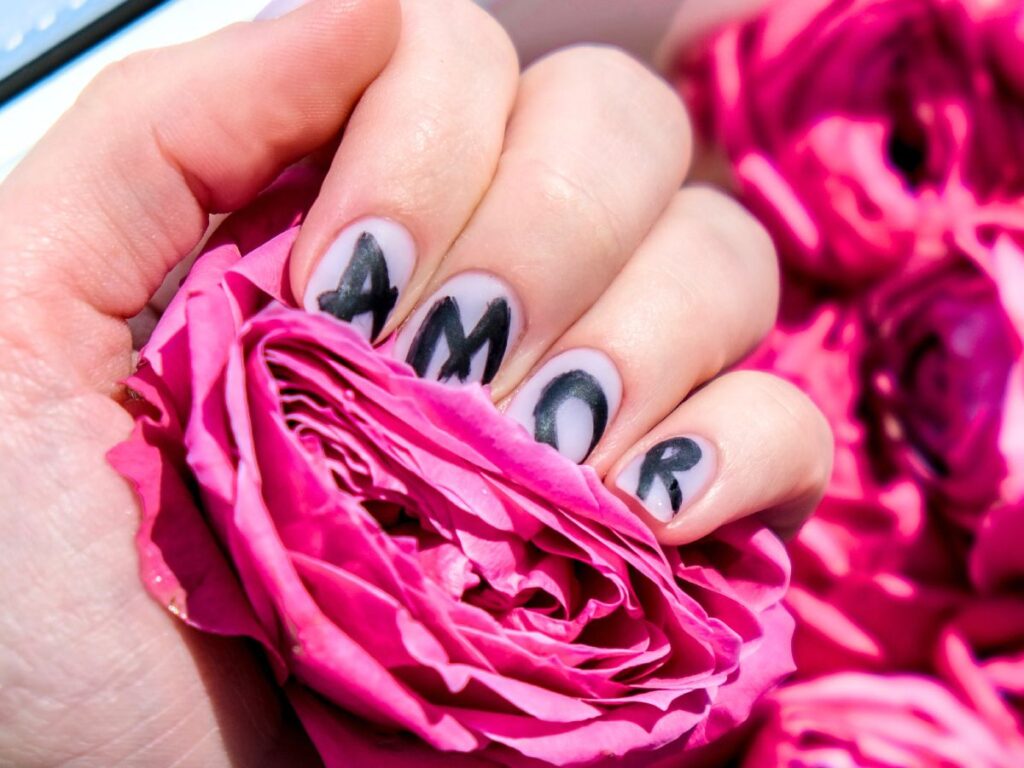 White nails with the word "AMOR" written letter by letter across four nails. Hand holding pink flower.