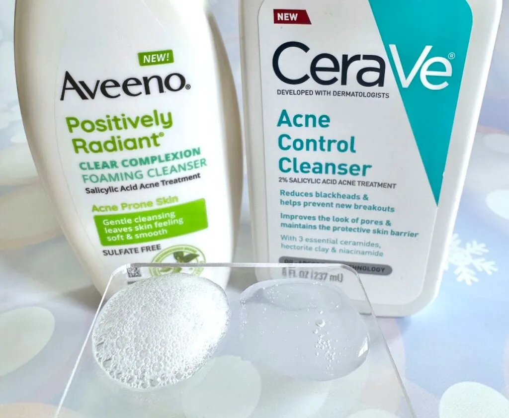 Aveeno Positively Radiant Clear Complexion Foaming Cleanser and CeraVe Acne Control Cleanser behind samples on clear spatula.