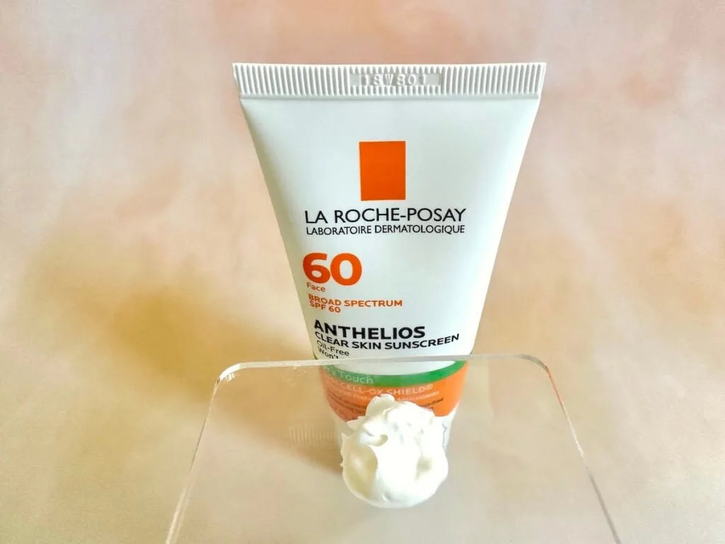 La Roche-Posay Anthelios Clear Skin Sunscreen SPF 60, tube behind sunscreen sampled on clear cosmetic spatula.