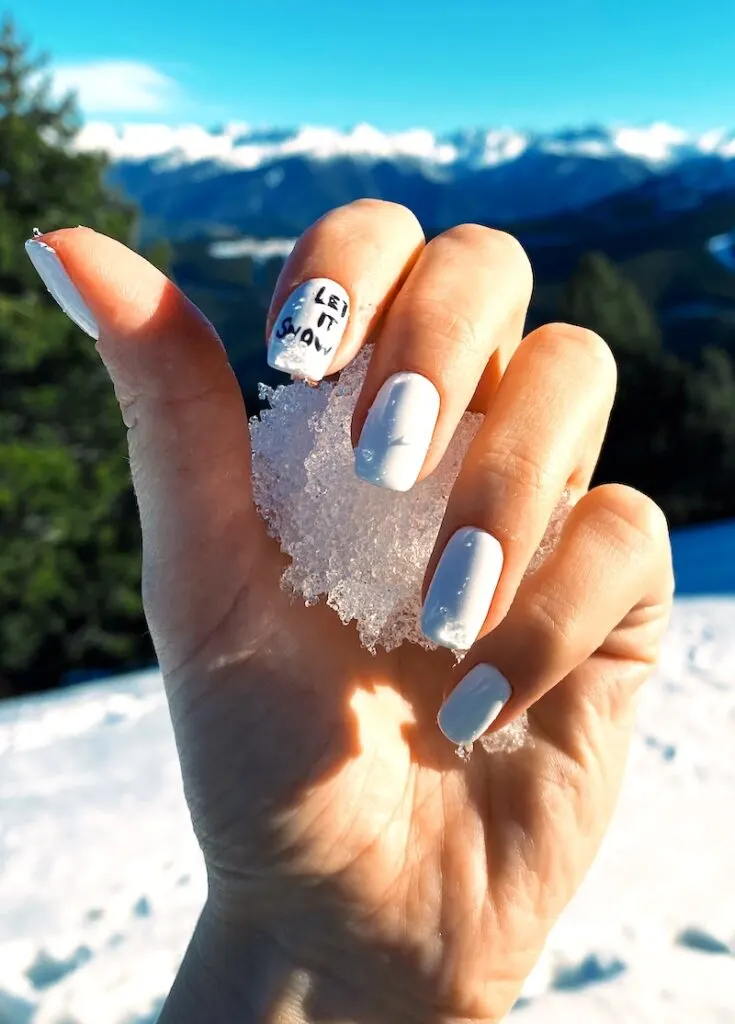 Let It Snow nail art on white nails holding a snowball outside on snow-covered bright beauitful day with blue sky.