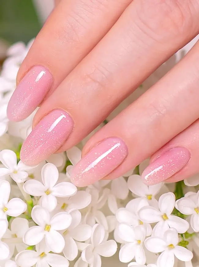 Golden glitter on oval pink nails touching white flowers.