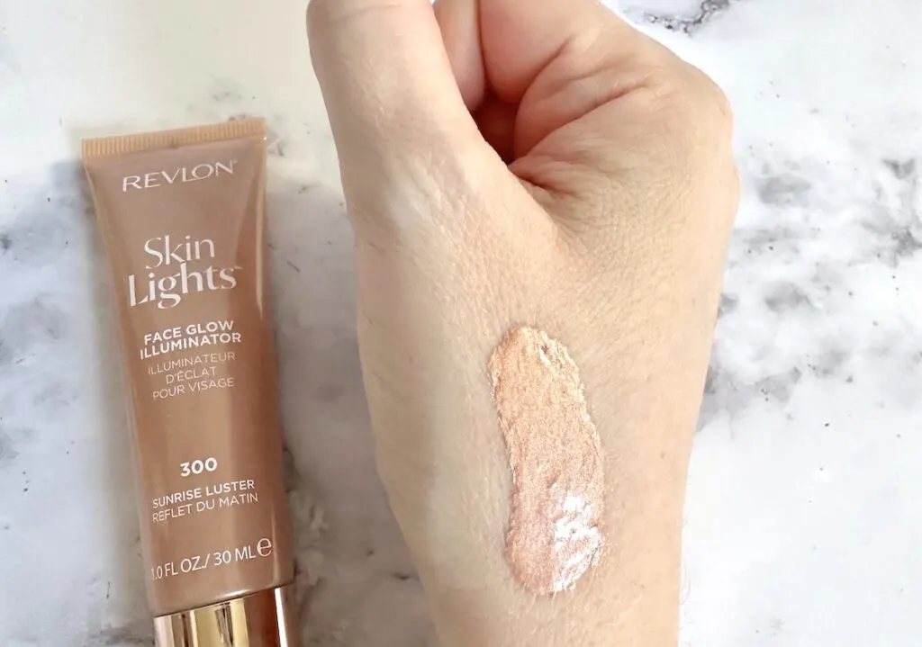 Revlon Skinlights Face Glow Illuminator in shade 300 Sunrise Luster. Flatlay of tube next to sample on hand.