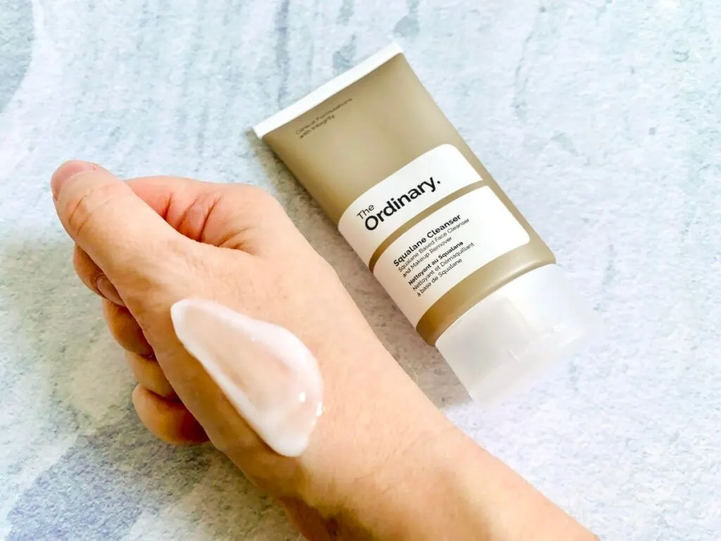 The Ordinary Squalane Cleanser tube and sampled on hand on a blue background.