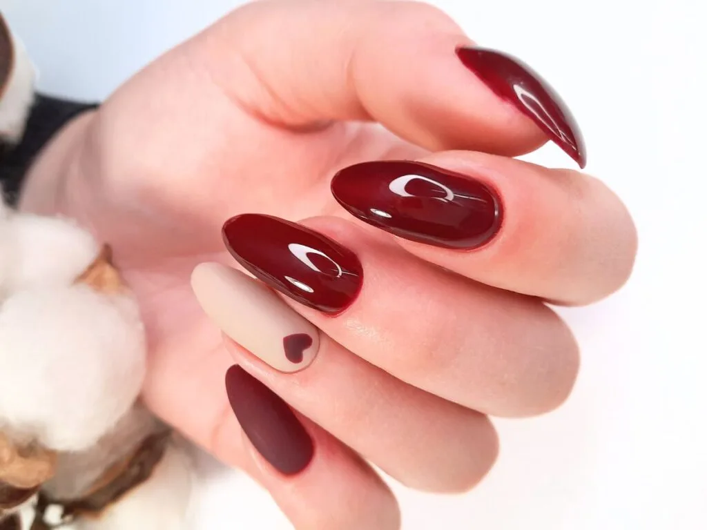 Deep burgundy long almond nails with one red heart on a neutral nail on the ring finger.