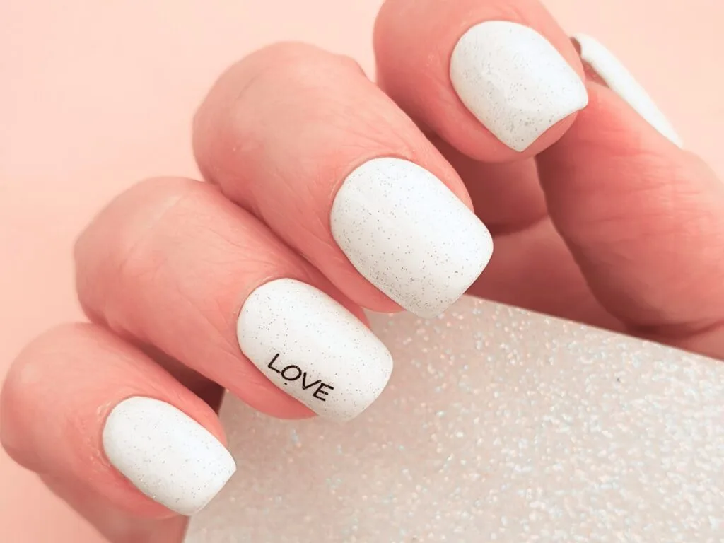 Matte white polish Valentines nails speckled with subtle grey flecks, with the ring finger displaying the word "LOVE" along the outer length of the nail.